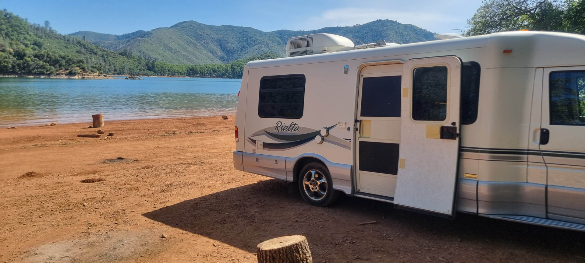 A small Class B motorhome parked on the red dirt shoreline at Beehive Point, Shasta Lake, with green hills across the water