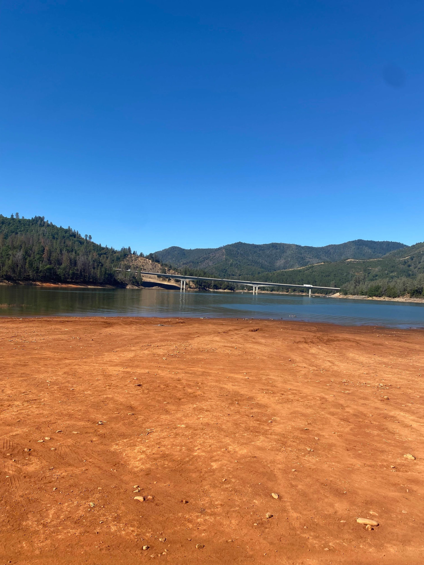 Wide red dirt beach at Gregory Beach on Shasta Lake with the highway bridge crossing the water beyond